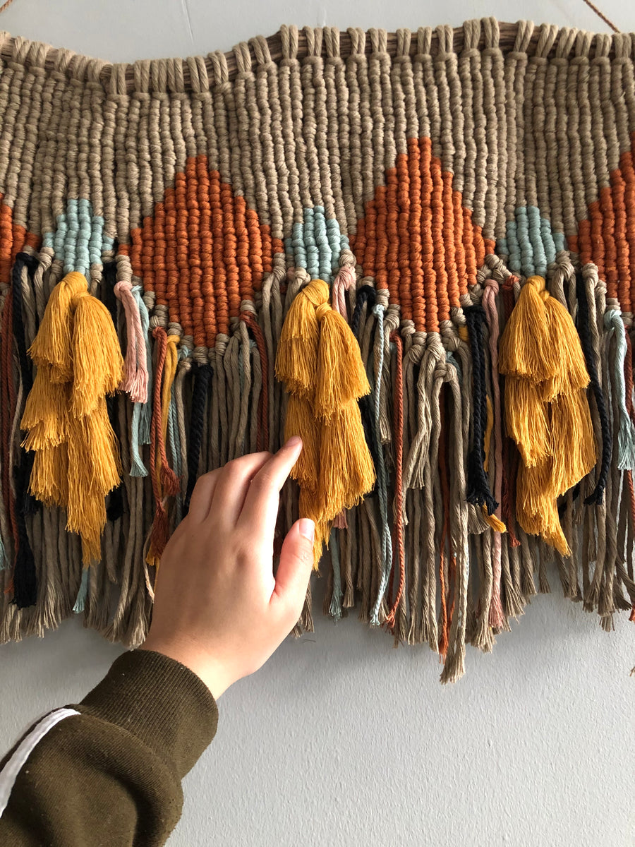 A hand touching a macrame hanging made of orange, light teal, mustard yellow, brown and black cords.