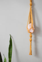 Load image into Gallery viewer, A yellow macrame hanger with spirals and a tassel holding a large cut rose quartz crystal inside.
