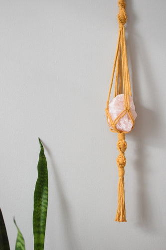 A yellow macrame hanger with spirals and a tassel holding a large cut rose quartz crystal inside.