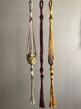 Load image into Gallery viewer, A trio of macrame hangers in beige, wine, and yellow. holding a ceramic skull and an amethyst crystal.
